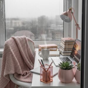 Pink desk lamp and laptop on a white desk with a view of rain through the window.