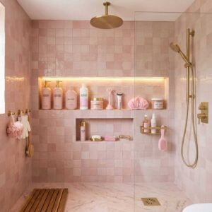 Modern walk-in shower featuring glossy blush pink square wall tiles, built-in storage niches, a premium matte gold rain shower head, and matching aesthetic shampoo dispensers.