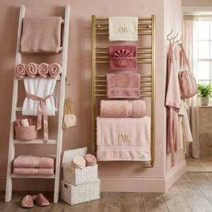 Brushed gold wall-mounted heated towel rail displaying monogrammed pink luxury bath towels next to a white wooden blanket towel storage ladder unit.