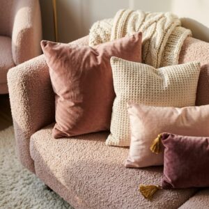 Close-up styling shot of a blush pink boucle couch layered with a mix of velvet, waffle knit, and silk throw pillows in terracotta, cream, and plum tones.