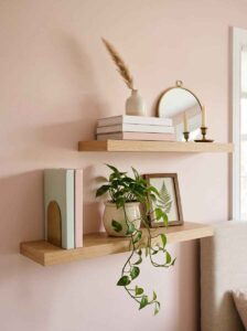 Natural oak floating shelves with minimalist blush decor, stacked design books, a trailing green pothos plant, and a small round gold mirror.
