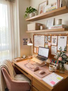 An organized college dorm desk setup featuring a pink laptop stand, mechanical keyboard, mouse, a large cork board with a calendar, motivation prints, and wooden shelves above.