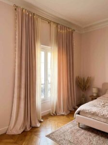 Floor-to-ceiling blush pink velvet curtains hung high on a gold rod to create the illusion of tall walls and a larger window frame in a minimalist bedroom.