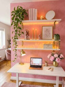 Styled white floating shelves with warm under-cabinet LED lighting, trailing pothos plant, books, and gold decor above a white desk setup.