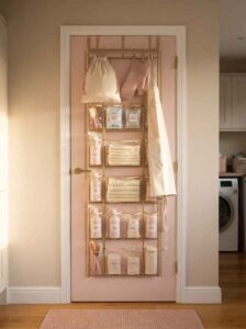 Over the door organizer with clear pockets on a blush pink door holding laundry sprays and cleaning supplies.