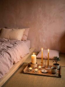 Relaxing low-light bedroom mood featuring burning pillar candles arranged on a rustic wooden floor tray beside a low-profile linen bed.
