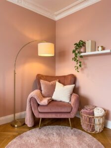 A beautifully styled cozy reading corner bedroom layout showcasing a rose velvet wingback chair, gold arc floor lamp, and a white floating wall shelf.