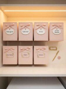 Pink fabric laundry storage bins with ribbon bows and ceramic labels for detergent, pods, and softener.