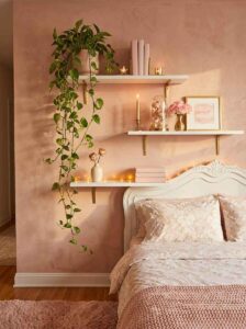 White floating shelves installed on a limewash pink wall above a bed, beautifully styled with pink books, candles, and a long trailing pothos ivy plant.