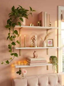Vertical storage solution for small rooms showing white floating shelves decorated with a trailing pothos plant, aesthetic pink books, gold candlesticks, and minimalist gold elephant bookends.