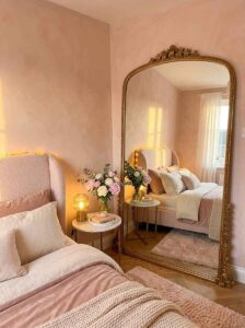 Small bedroom mirror placement featuring a giant arched gold floor mirror reflecting warm afternoon sunlight, a tufted pink bed frame, and chic marble bedside tables.