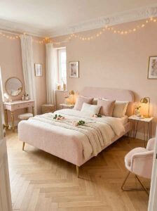 Small bedroom furniture arrangement featuring a light blush upholstered bed frame with slim gold legs, a matching pink vanity desk, chevron wood flooring, and delicate string lights.