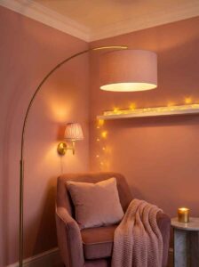 Atmospheric warm lighting bedroom reading nook aesthetic created by a gold arc floor lamp, a matching wall sconce, and subtle string fairy lights over a pink chair.