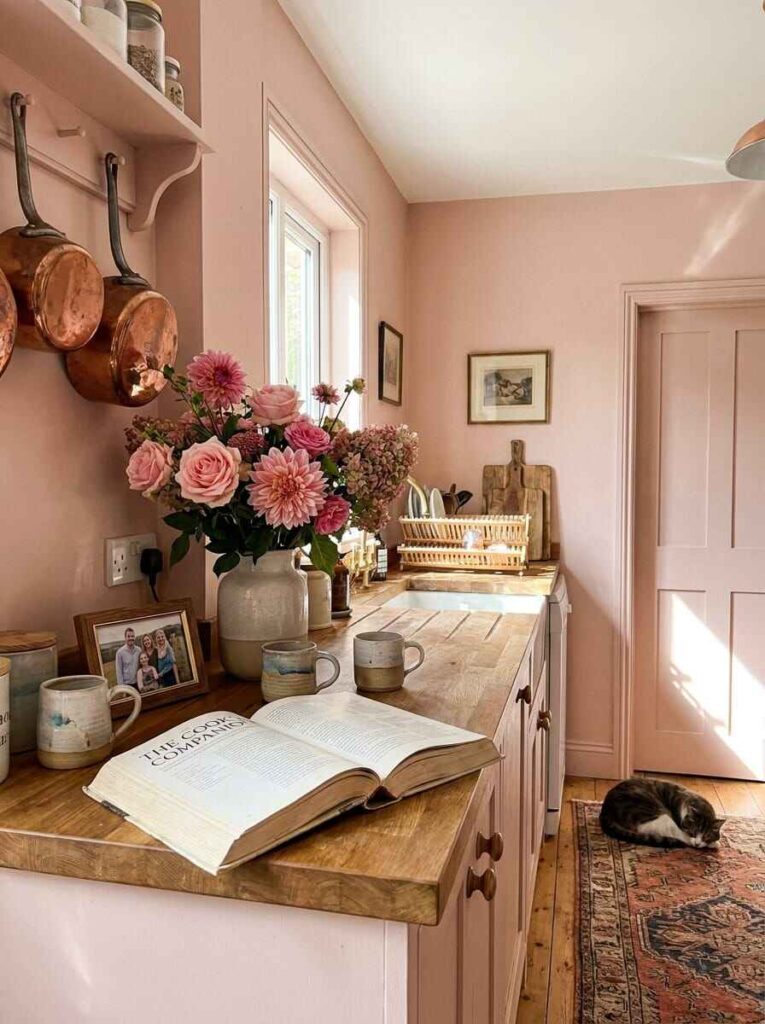 Cozy wooden kitchen counter with an open cookbook, family photo frame, handmade pottery mugs, copper pans, and pink dahlias.